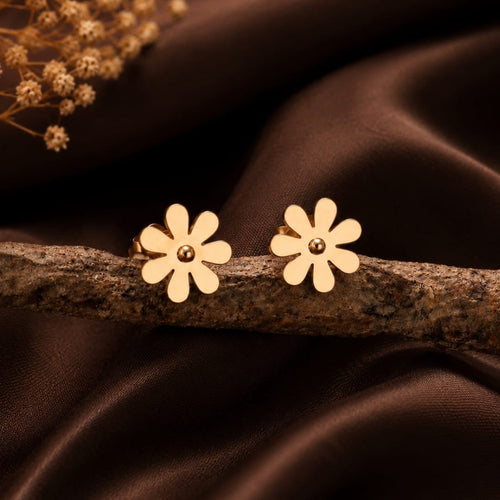 A pair of gold daisy flower stud earrings with a small central bead, resting on a textured branch over a draped dark brown satin fabric background with dried flowers in the corner.